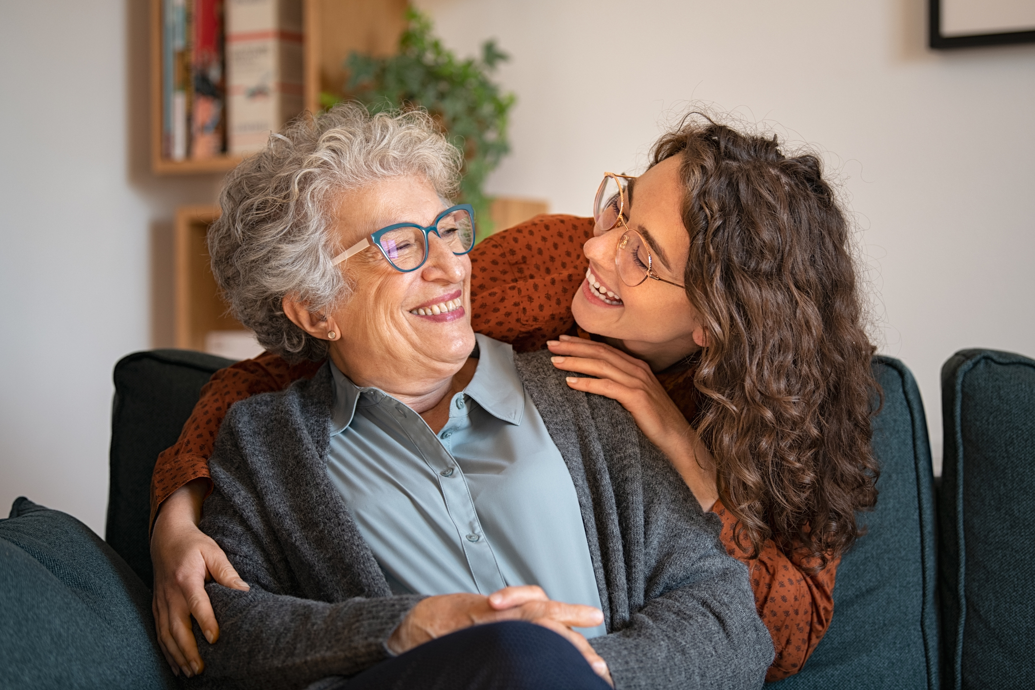 Old grandmother and adult granddaughter hugging at home and looking at each other. Happy senior mother and young daughter embracing with love on sofa. Happy young woman hugging from behind grandma with love.