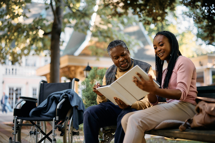 Happy African American woman looking at photos in family album with her senior father while vising him at nursing home.