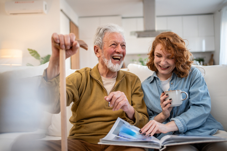 Senior man using a walking cane sits on the sofa with a young woman, both laughing and enjoying tea while looking through a photo album, sharing cherished memories and moments of joy