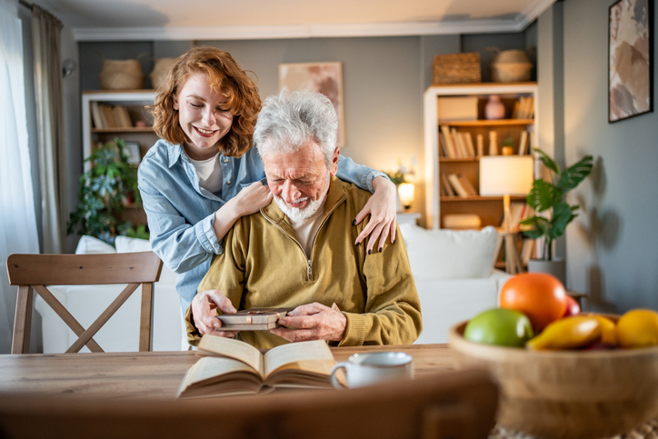 Granddaughter hugging her grandfather while he reads a book and shares a picture, creating a warm moment of connection and joy in the cozy living room at home